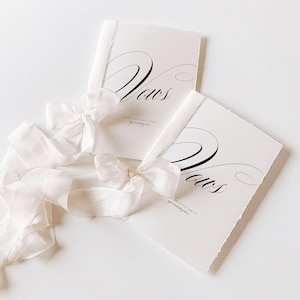 May include: Two white vow books with the word "Vows" in elegant black script. Each book is tied with a delicate white ribbon. The books have a deckled edge and the text "to my beloved, entwined, my wedding day."