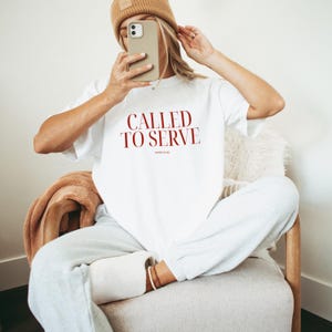 May include: A white t-shirt with the text "CALLED TO SERVE" in red lettering. The shirt is being worn by a person sitting on a chair.