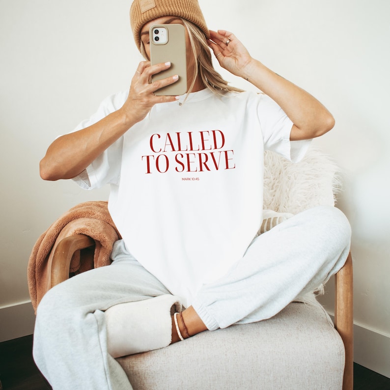 May include: A white t-shirt with the text "CALLED TO SERVE" in red lettering. The shirt is being worn by a person sitting on a chair.