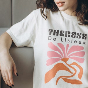 以下が含まれることがあります： 白いTシャツに、ピンクとオレンジの花のグラフィックと黒い文字で「Therese De Lisieux」と書かれています。