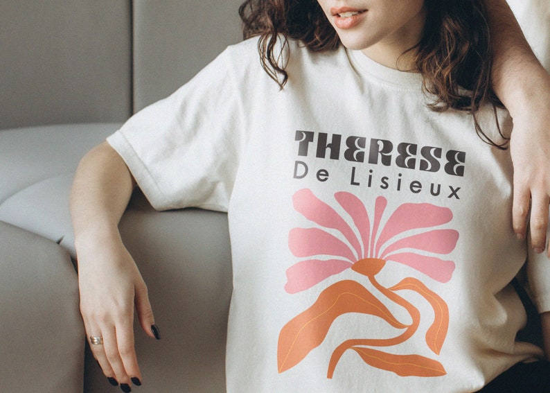 以下が含まれることがあります： 白いTシャツに、ピンクとオレンジの花のグラフィックと黒い文字で「Therese De Lisieux」と書かれています。