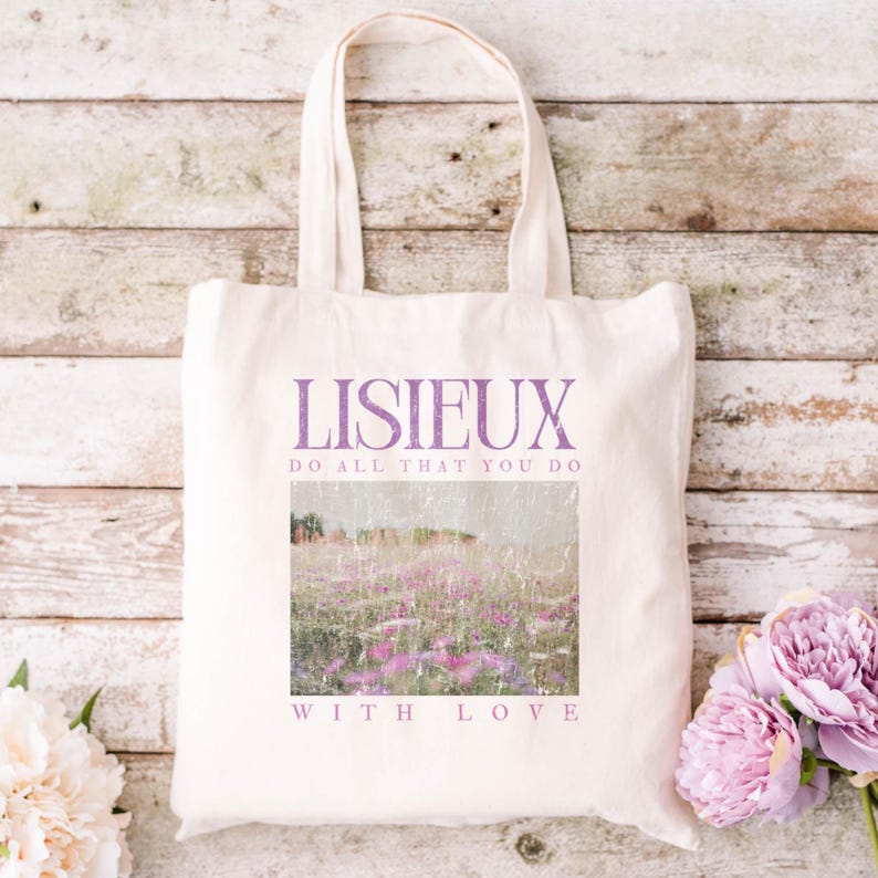 May include: Cream-colored tote bag with the word "LISIEUX" in purple, along with the phrases "DO ALL THAT YOU DO" and "WITH LOVE." The bag features a rectangular image of a field of purple flowers. The bag has two handles.