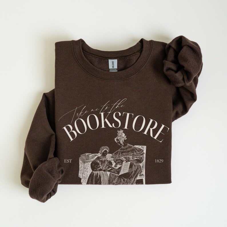 May include: Brown sweatshirt with a white graphic that says "Take me to the bookstore" with an illustration of two women reading a book. The text "EST 1829" is also included in the graphic.