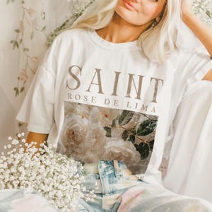 Op de afbeelding: Wit T-shirt met een bruine grafische print van bloemen en de tekst "SAINT ROSE DE LIMA".