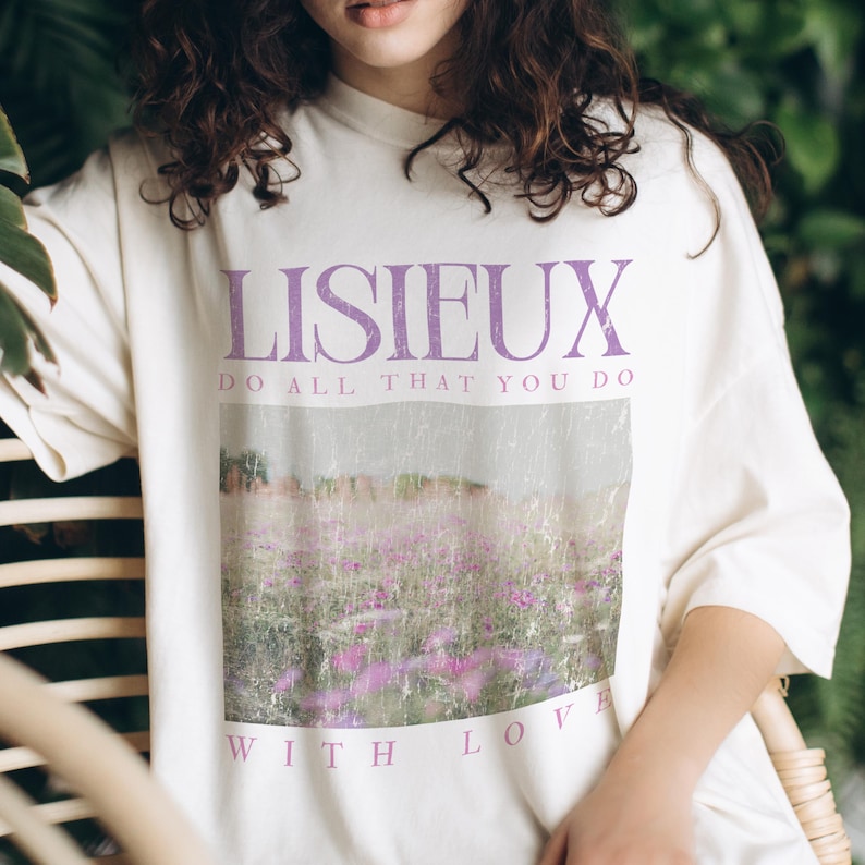 May include: Cream-colored t-shirt with the word "LISIEUX" in purple, above the text "DO ALL THAT YOU DO." A faded image of a field of purple flowers is below the text, with the words "WITH LOVE" at the bottom.