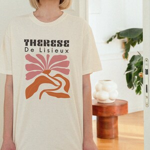 以下が含まれることがあります： クリーム色のTシャツに、ピンクとオレンジのスタイリッシュな花をモチーフにしたグラフィックデザインが施されています。Tシャツには黒い文字で「Therese De Lisieux」と印刷されています。