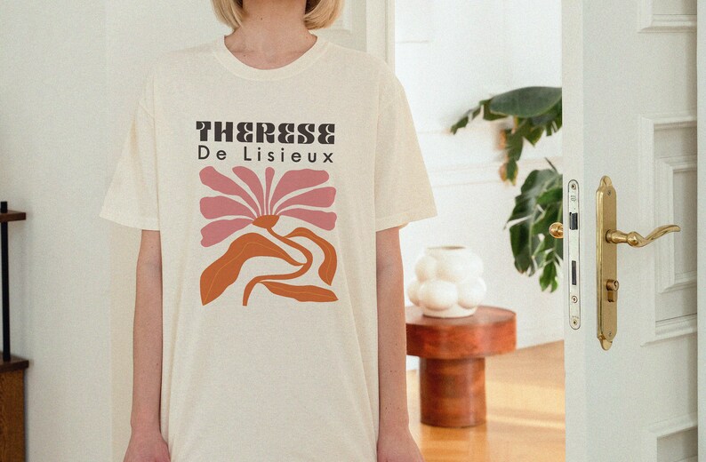 以下が含まれることがあります： クリーム色のTシャツに、ピンクとオレンジのスタイリッシュな花をモチーフにしたグラフィックデザインが施されています。Tシャツには黒い文字で「Therese De Lisieux」と印刷されています。