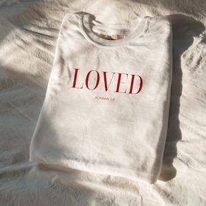 May include: White t-shirt with the word "LOVED" in red lettering and the text "ROMANS 5:8" underneath.