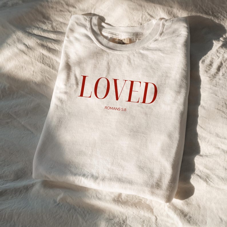 May include: White t-shirt with the word "LOVED" in red lettering and the text "ROMANS 5:8" underneath.