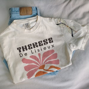 以下が含まれることがあります： 「Therese De Lisieux」とピンクとオレンジの花の模様が書かれたブラウンのグラフィックプリントが施された白いTシャツです。