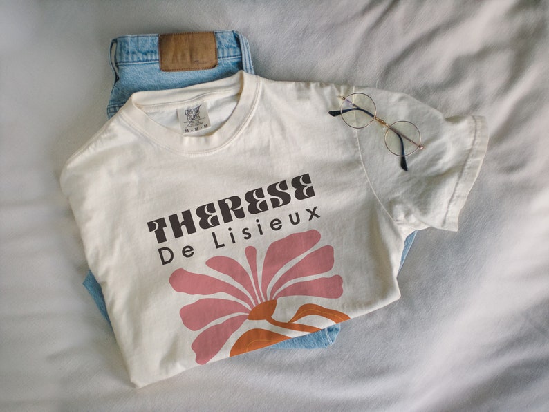 以下が含まれることがあります： 「Therese De Lisieux」とピンクとオレンジの花の模様が書かれたブラウンのグラフィックプリントが施された白いTシャツです。
