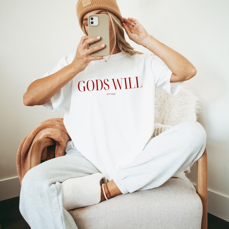 May include: White t-shirt with the text "GODS WILL" in red lettering. The shirt is being worn by a person sitting on a chair.