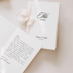 May include: A pair of white vow books tied with a light pink ribbon. The cover of one book reads "His Vows" in elegant script, with the names "Emma & Noah" and a date below. The other book displays handwritten vows.