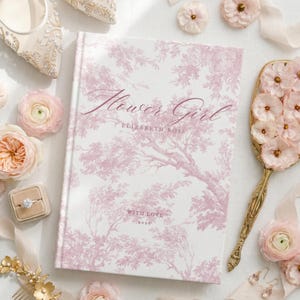 May include: A hardcover book with a white cover featuring a pink toile de jouy tree pattern. The text reads Flower Girl, Elizabeth Rose, and With Love 2026. The book is surrounded by pink flowers, a gold hand mirror, a ring in a box, and jewelry.