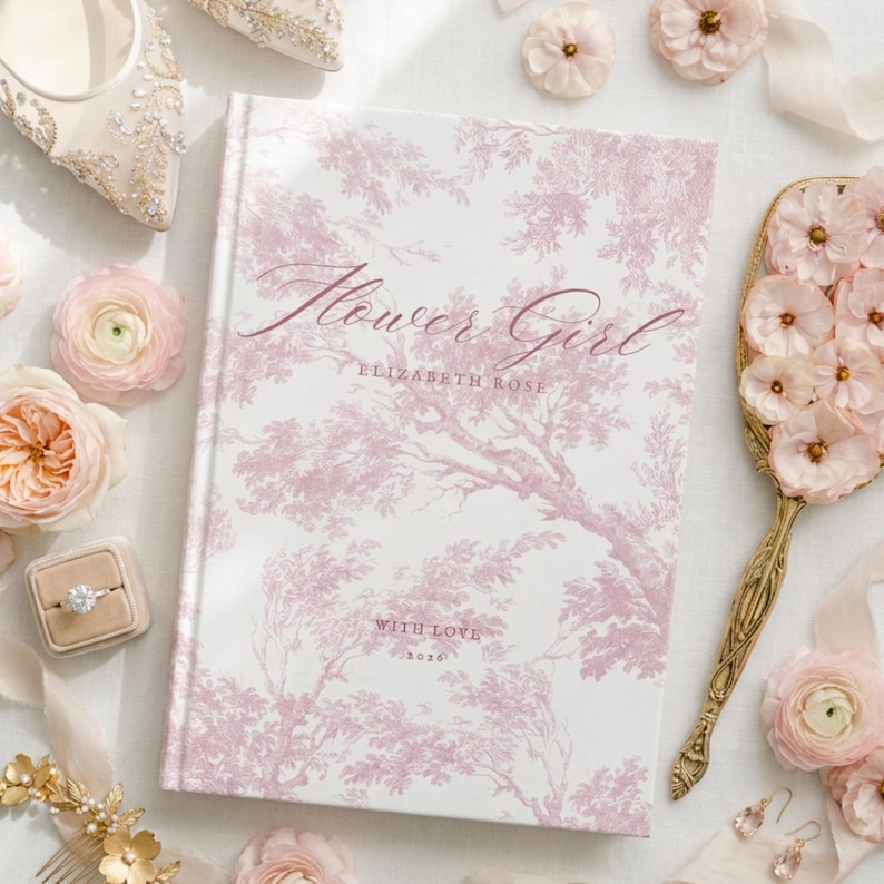 May include: A hardcover book with a white cover featuring a pink toile de jouy tree pattern. The text reads Flower Girl, Elizabeth Rose, and With Love 2026. The book is surrounded by pink flowers, a gold hand mirror, a ring in a box, and jewelry.