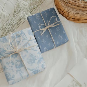 May include: Two wrapped gifts with blue floral patterned paper and twine bows. One gift is wrapped in a solid blue floral pattern, and the other in a light blue floral pattern. The gifts are on a white surface.
