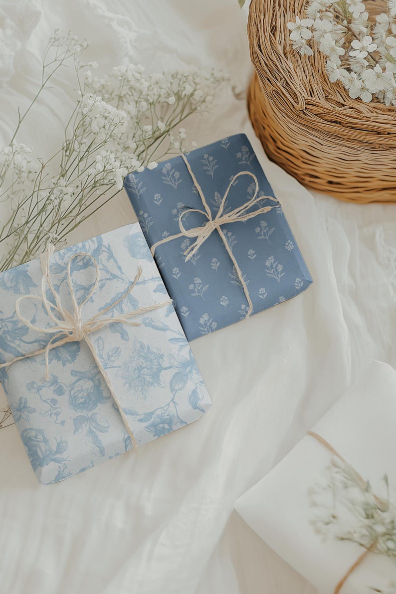 May include: Two wrapped gifts with blue floral patterned paper and twine bows. One gift is wrapped in a solid blue floral pattern, and the other in a light blue floral pattern. The gifts are on a white surface.