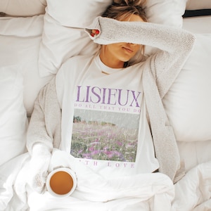 May include: A white t-shirt with the word "LISIEUX" in purple, along with the phrase "DO ALL THAT YOU DO" and "WITH LOVE". The shirt features a landscape image with purple flowers. A person is wearing the shirt and a gray cardigan, lying on a bed with a cup of tea.