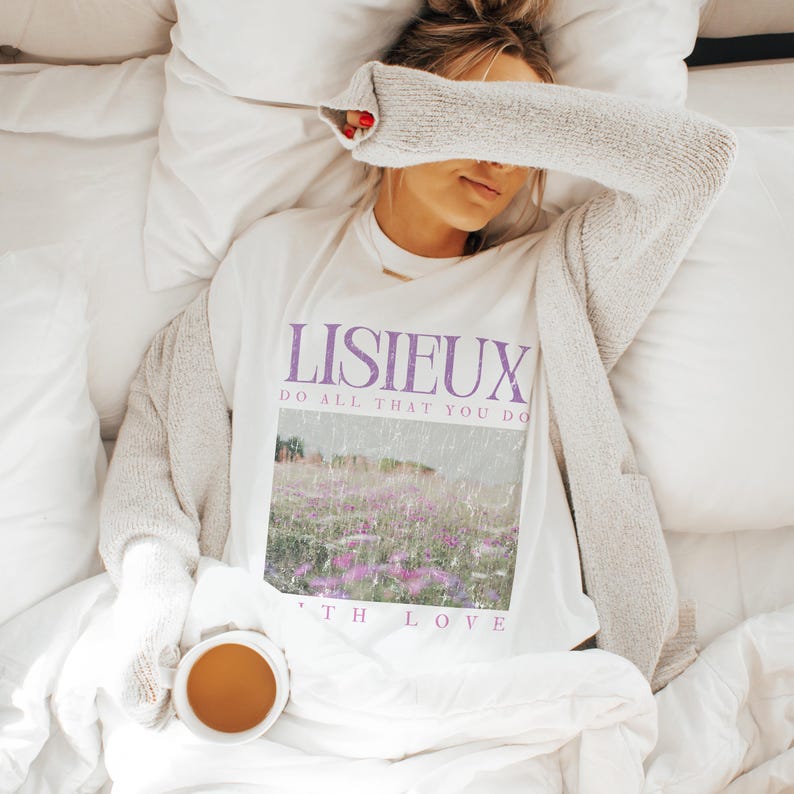 May include: A white t-shirt with the word "LISIEUX" in purple, along with the phrase "DO ALL THAT YOU DO" and "WITH LOVE". The shirt features a landscape image with purple flowers. A person is wearing the shirt and a gray cardigan, lying on a bed with a cup of tea.