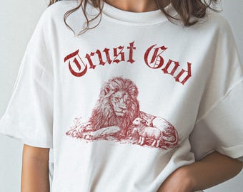 May include: White t-shirt with a red graphic of a lion and a lamb, and the words "Trust God" in a decorative font. The design is centered on the front of the shirt.