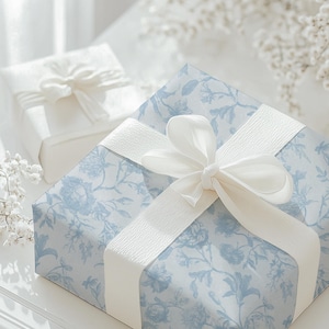 May include: Two wrapped gifts. The larger gift is wrapped in light blue floral patterned paper and tied with a white ribbon. A smaller gift, wrapped in white paper, is in the background. The gifts are on a white surface.