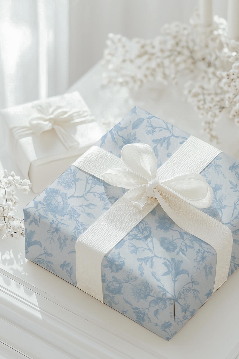 May include: Two wrapped gifts. The larger gift is wrapped in light blue floral patterned paper and tied with a white ribbon. A smaller gift, wrapped in white paper, is in the background. The gifts are on a white surface.