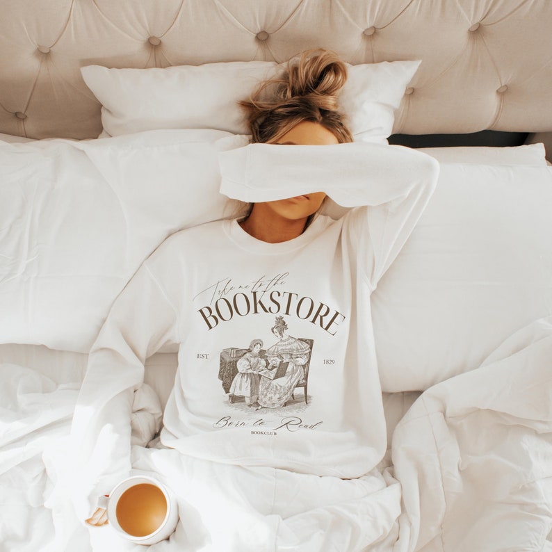 May include: A white sweatshirt with a brown graphic print that says "Take me to the Bookstore" with an illustration of a woman reading to a child. The text "Est. 1829" and "Born to Read Book Club" are also printed on the sweatshirt.