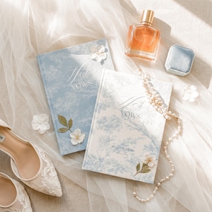 May include: Two hardcover vow books with blue and white toile patterns, labeled His Vows and Her Vows, accompanied by a glass perfume bottle, a small blue velvet ring box, a string of pearls, white fabric flowers, and white lace wedding shoes.