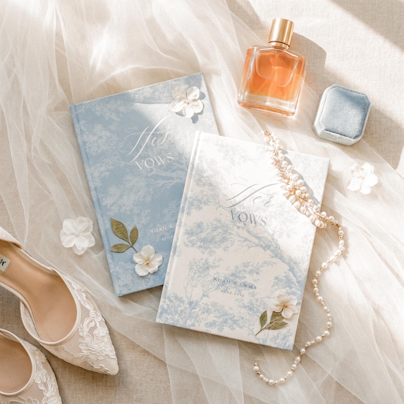 May include: Two hardcover vow books with blue and white toile patterns, labeled His Vows and Her Vows, accompanied by a glass perfume bottle, a small blue velvet ring box, a string of pearls, white fabric flowers, and white lace wedding shoes.
