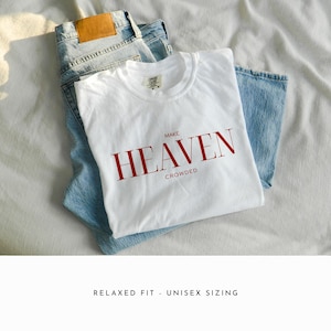 Op de afbeelding: Wit T-shirt met rode tekst die "MAKE HEAVEN CROWDED" luidt. Het shirt is gevouwen en gelegd op een blauwe jeans.