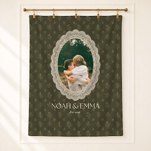 May include: A dark olive green wall hanging featuring a repeating floral pattern, a central oval photo of two people embracing framed by a white lace border, and the white text "NOAH & EMMA" followed by "Est. 2026" printed below the frame.