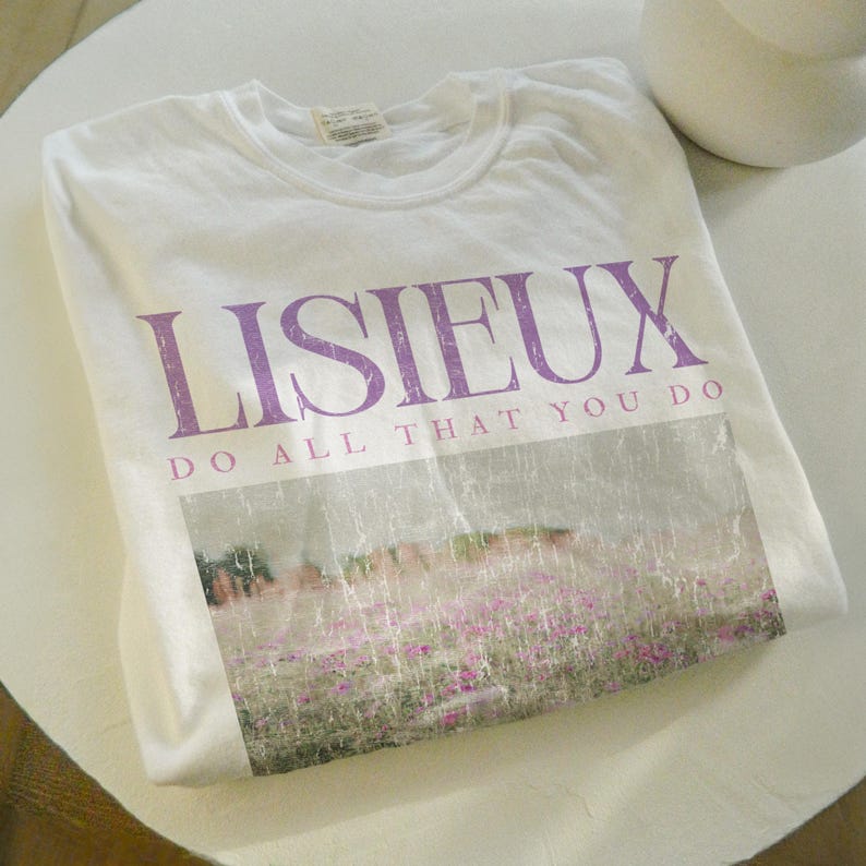 May include: A white t-shirt with the word "LISIEUX" in purple, above the phrase "DO ALL THAT YOU DO." The shirt features a faded image of a field of pink flowers. The shirt is folded on a white surface.