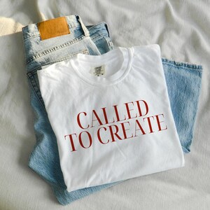 May include: White t-shirt with the text "CALLED TO CREATE" in red lettering. The t-shirt is folded and placed on top of a pair of blue jeans.