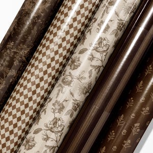May include: An assortment of rolled wrapping paper with various patterns. Designs include a brown and cream checkered pattern, floral prints, and a dark brown toile pattern. The paper is glossy and appears to be new.