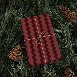 May include: A wrapped gift with a burgundy striped pattern, tied with twine, rests on a bed of green pine needles and pine cones. The gift box is rectangular and the twine is tied in a bow.