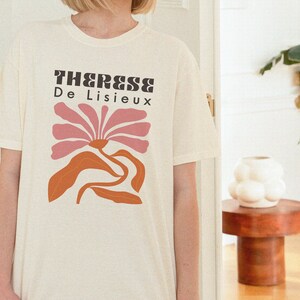 以下が含まれることがあります： ピンクの花と黒い文字「Therese De Lisieux」がプリントされた白いTシャツです。