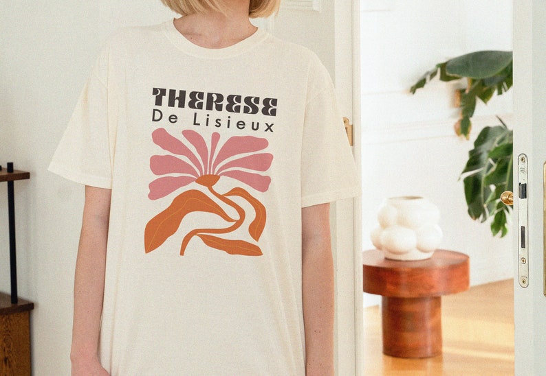 以下が含まれることがあります： ピンクの花と黒い文字「Therese De Lisieux」がプリントされた白いTシャツです。