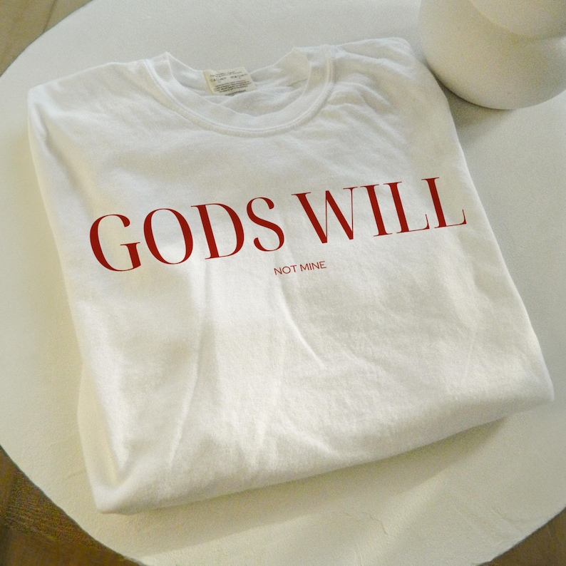 May include: White t-shirt with the text "GODS WILL NOT MINE" printed in red.