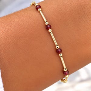 Könnte beinhalten: Ein zartes Armband mit abwechselnd kleinen goldfarbenen Kugeln und längeren goldfarbenen zylindrischen Stäbchen, durchsetzt mit tiefroten facettierten Edelsteinperlen. Das Design erzeugt ein wiederkehrendes Muster aus Metall und dunklem Stein.