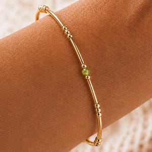 May include: A thin gold bangle bracelet featuring a series of small, polished gold beads and a single, central faceted peridot green gemstone. The bracelet has a smooth, metallic finish and a minimalist design that encircles the wrist.