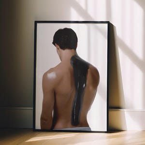May include: A framed art print featuring a person's back with a dark, abstract design. The print is in a black frame, leaning against a white wall. The room has wooden floors and natural light.
