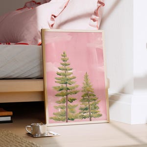 Op de afbeelding: Een ingelijste kunstdruk met twee groene dennenbomen tegen een roze lucht. Het kunstwerk staat op een houten vloer, leunend tegen een witte muur. Een klein kopje en een geweven mat liggen op de voorgrond.