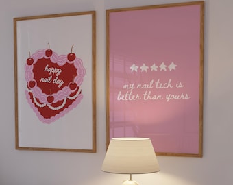 May include: Two framed prints with wooden frames hang on a white wall. One print features a pink cake design with cherries and the text "happy nail day." The other is pink with the text "my nail tech is better than yours." A white lamp sits on a white cabinet.