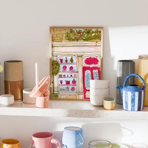 May include: A collection of colorful ceramics and decorative items displayed on white shelves. Items include mugs, bowls, a teapot, a framed illustration of a shop, and various vases. The color palette includes blue, yellow, pink, and orange.