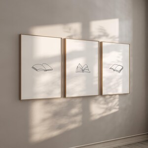 Puede incluir: Tres impresiones artísticas enmarcadas con marcos dorados, cada una con un dibujo lineal minimalista de libros abiertos sobre un fondo blanco. Los dibujos negros varían en el grado de apertura de los libros.