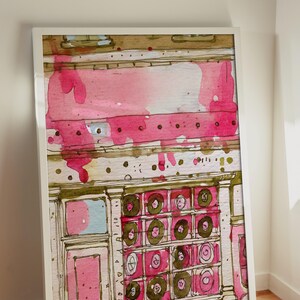 May include: Watercolor painting of a building facade with pink and brown accents. The artwork features a white frame and is leaning against a white wall and wooden floor. The building has a series of circular designs.