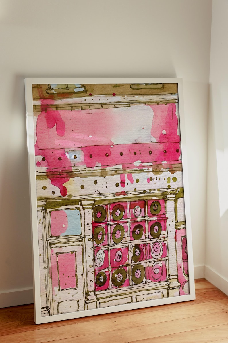 May include: Watercolor painting of a building facade with pink and brown accents. The artwork features a white frame and is leaning against a white wall and wooden floor. The building has a series of circular designs.