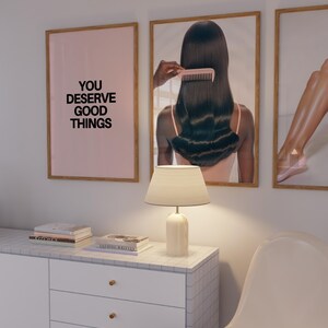 May include: A room interior featuring three framed art prints. One print has the text "YOU DESERVE GOOD THINGS" on a pink background. A white lamp sits on a white cabinet with gold knobs. A white chair is in the foreground.