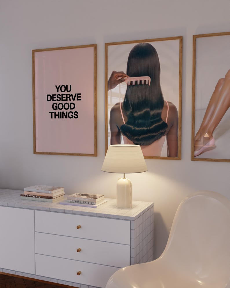 May include: A room interior featuring three framed art prints. One print has the text "YOU DESERVE GOOD THINGS" on a pink background. A white lamp sits on a white cabinet with gold knobs. A white chair is in the foreground.