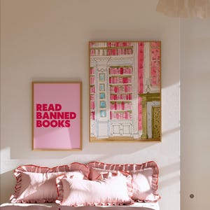 May include: A bedroom scene featuring a bed with pink bedding and pillows with red trim. Two framed artworks hang on the wall: one with the text "READ BANNED BOOKS" and another depicting a library interior. The room is painted in soft pink tones.